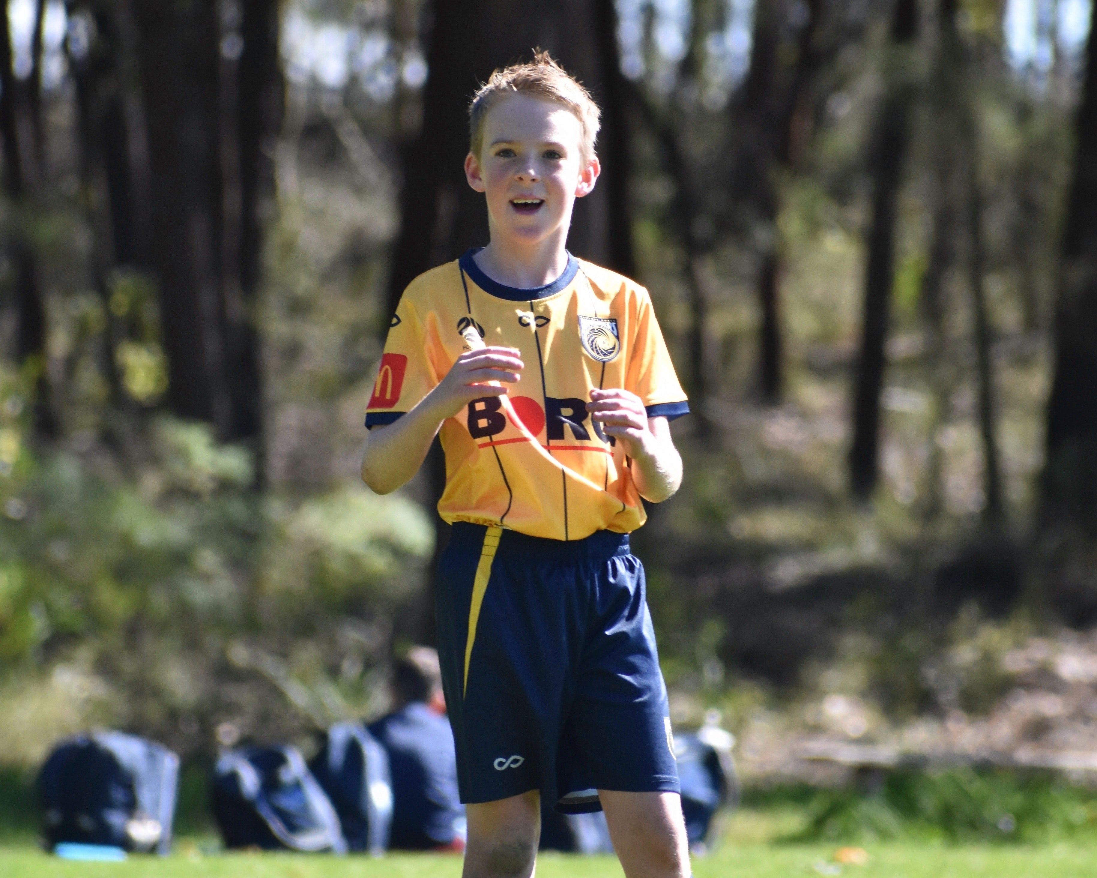 Young boy in sports uniform holding a microphone outdoors with trees in the background